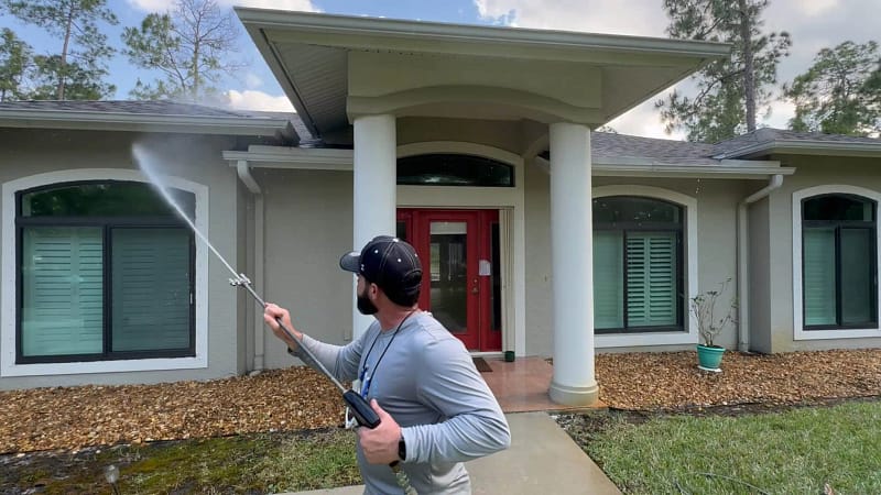 "Technician performing soft wash on the exterior of a modern house with white columns and red front door"